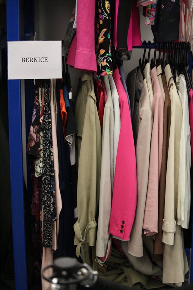 A metal clothing rack full of some clothes in various different colours including bright pink and black next to a square white sign that has Bernice written in bold black writing on it, on a dark background.