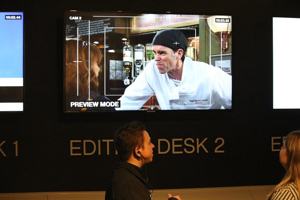 A screen in a dark room that has a picture of an angry man wearing a white chef's outfit in a brightly lit Pub on it and Edit Desk written in bold white writing below it, on a light background.