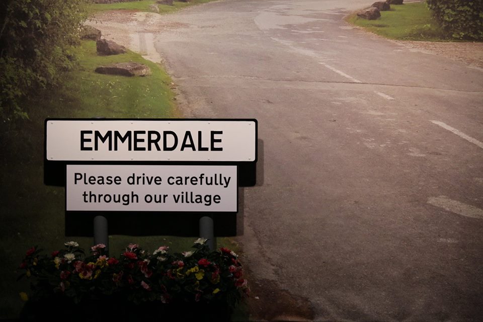 A white rectangular sign that has Emmerdale written in bold black capital writing on it next to another white rectangular sign that has please drive carefully through our village written in bold black writing and a picture of some shrubs on it, on a grey background.