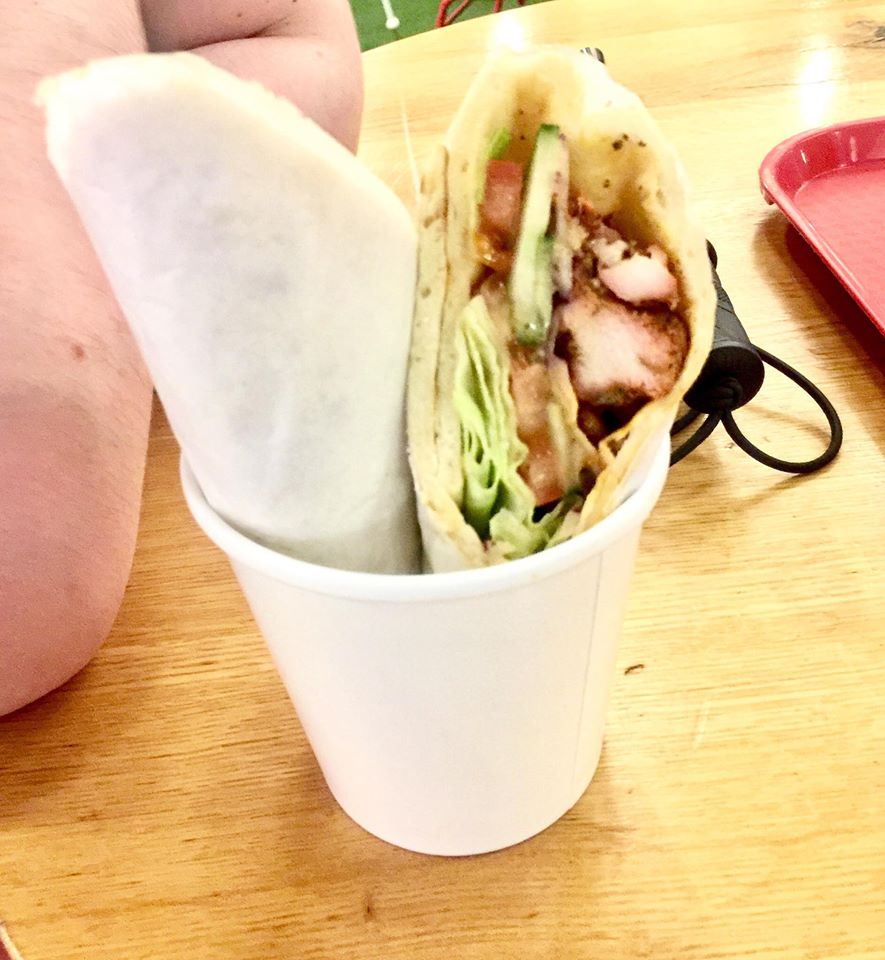 A wrapped up Flatbread filled with some dark Red pieces of Chicken Breast, some light Green pieces of Lettuce, and some dark Green pieces of Cucumber inside a bright white polystyrene cup on a light wooden table, on a light background.