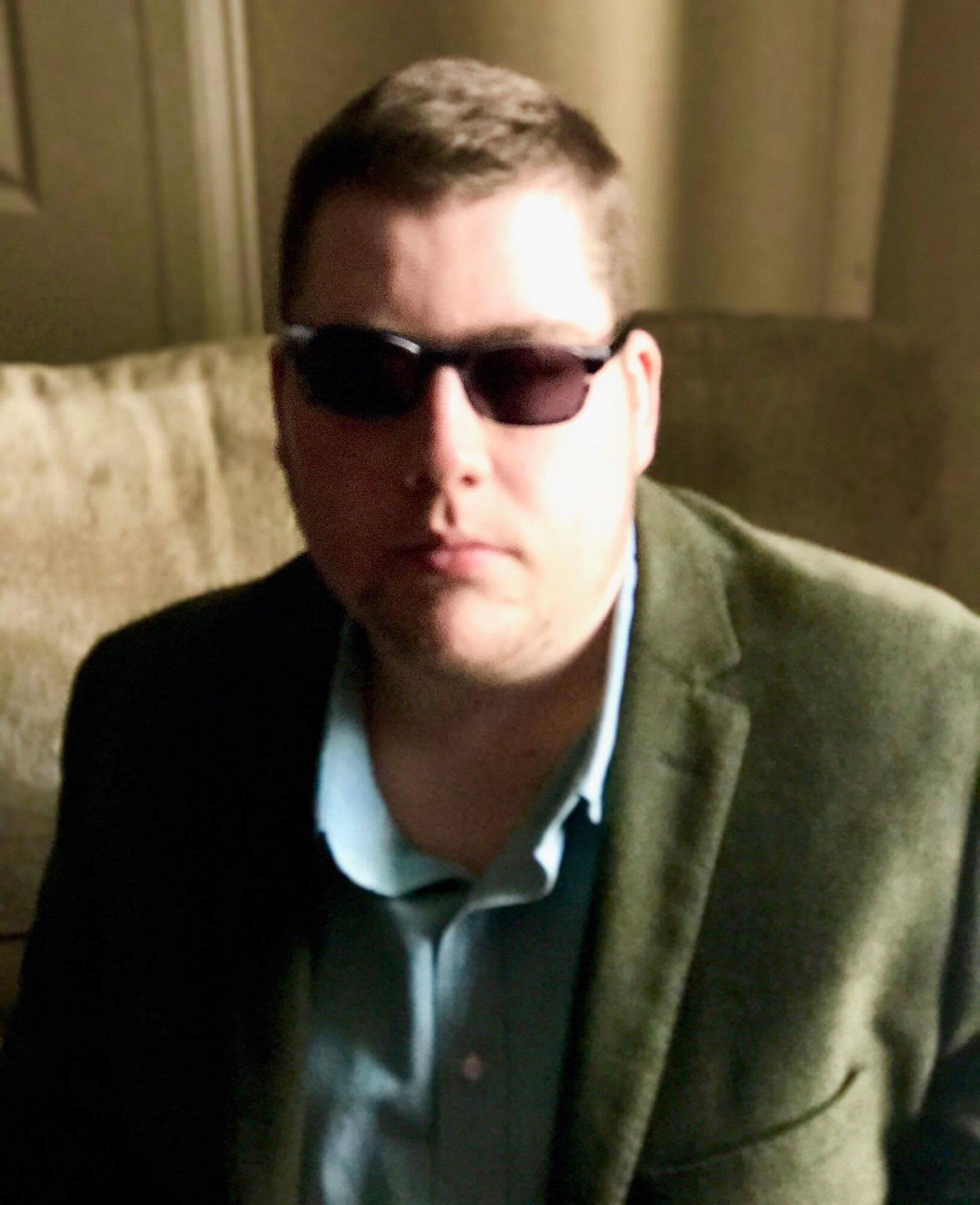 Me wearing a green blazer, a light green shirt, and a pair of black thin plastic Sunglasses with Grey lenses, on a light background.