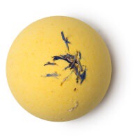 A bright yellow coloured spherical bath bomb with some blue cornflowers sprinkled on top of it, on a white background.