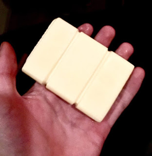 A bright white coloured rectangular massage bar with some indentations in it to make it look like a small chocolate bar on a light pink coloured hand, on a light background.