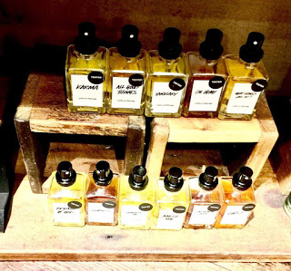Two rows of five rectangular clear glass fragrance bottles with some white labels that have the names of the fragrances written on them in bold black writing on a light wooden shelf, on a light background.