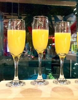 Some tall clear glass champagne flutes full of some bright orange juice on a clear glass table, on a bright background.