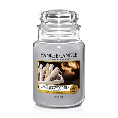 A tall glass jar full of some light blue coloured wax with a label that has yankee candle written in white writing, Crackling Wood Fire written in black writing, and a picture of some light blue coloured wood in a square pot next to a roaring wood fire on it, on a white background.