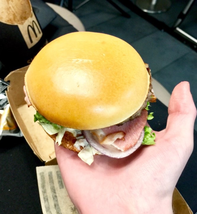 A pale Pink hand holding a A golden Brioche style Burger Bun with a brown Burger Patty, some white Coleslaw, and some brown Barbecue Sauce inside it, on a white background.
