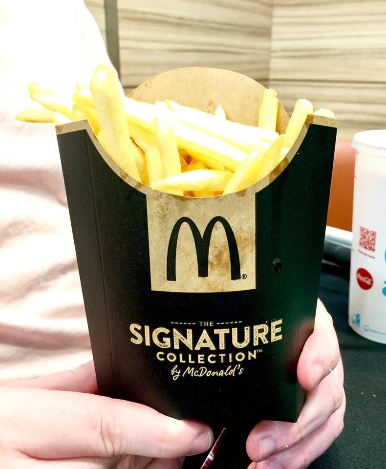 Some golden crispy Fries inside a Black semi-circled cardboard carton that has a black McDonald's logo inside a gold square and The Signature Collection written in silver writing on it, on a white background.