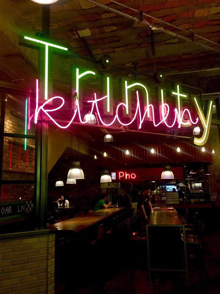 A large wide room full of chairs with a light above it that has Trinity written in bright white writing and kitchen written in bright pink cursive writing on it with some spotlights in the background, on a dark background.