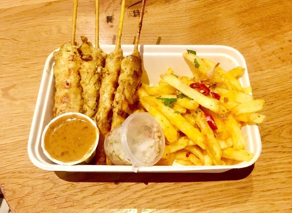 Some pieces of bright golden Chicken on some light brown skewers next to a pile of thin Golden Fries sprinkled with some bright red chilli flakes and light green spring onions, a white circular tub full of some golden Satay dip, and a clear circular tub full of some bright green Shallots inside a white rectangular take away box on a golden table, on a bright background.