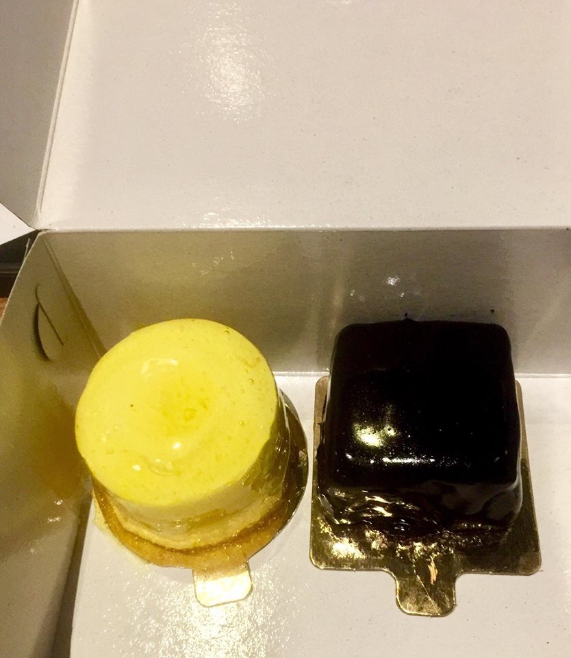 A bright cylindrical white chocolate sponge cake covered in some bright yellow lemon curd next to a square dark brown chocolate cake covered in some dark brown ganache inside a white rectangular box, on a bright background.