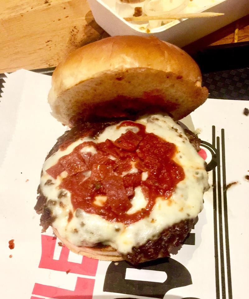 A brioche Bun filled with a dark brown beef patty, some pale golden melted Cheese and some bright red Tomato Sauce on a white and red patterned piece of paper, on a light background.