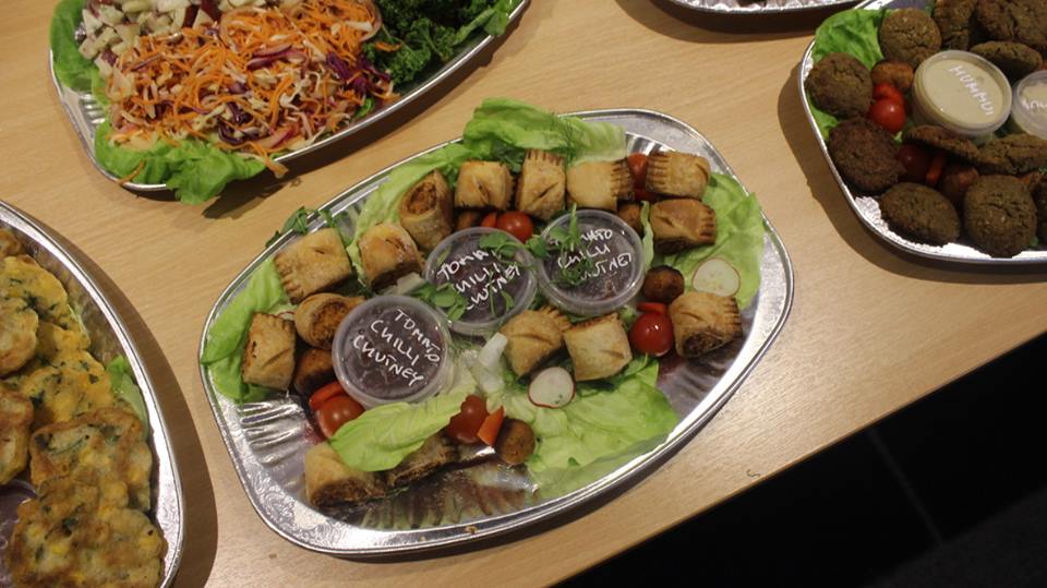 A large silver oval plate with some triangular brown bread sandwiches and some bright green apples on it on a light wooden table, on a light background.