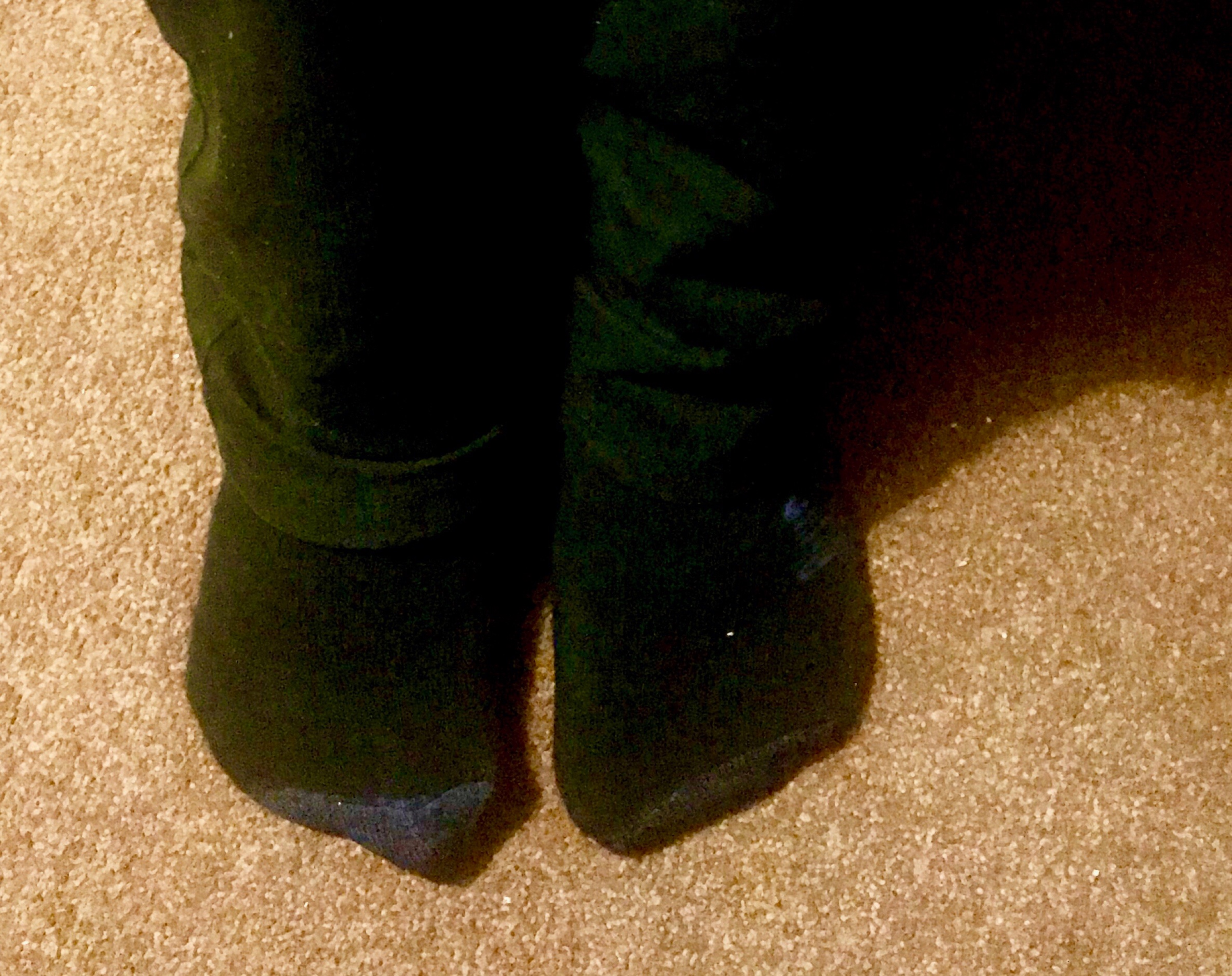 two feet wearing a pair of long black socks with grey tips on a cream carpet, on a light background.