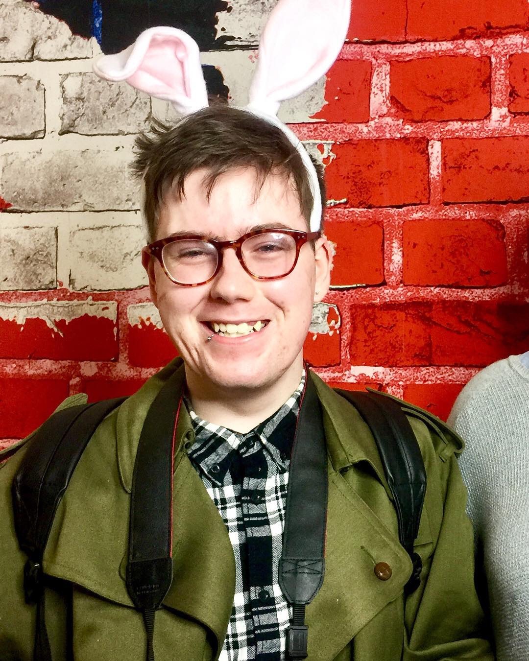A person smiling with messy dark brown short hair wearing a pair of white bunny ears, a grey and white checked shirt, and a grey overcoat, on a red brick background.