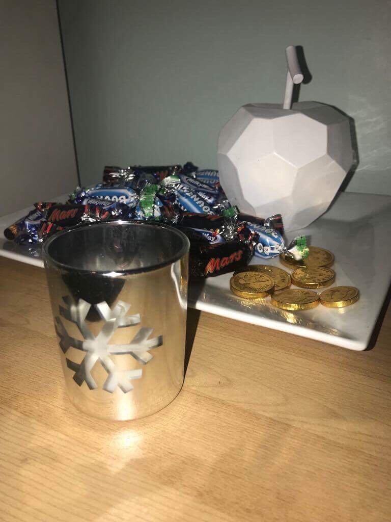 A rectangular white plate that has some chocolate coins in silver wrappers and a large white apple ornament on it on a large rectangular light wooden chest of draws, on a light background.