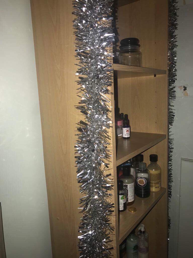 Some shiny silver tinsel wrapped around a tall light wooden rectangular bookcase filled with some various different shaped bottles, on a light background.