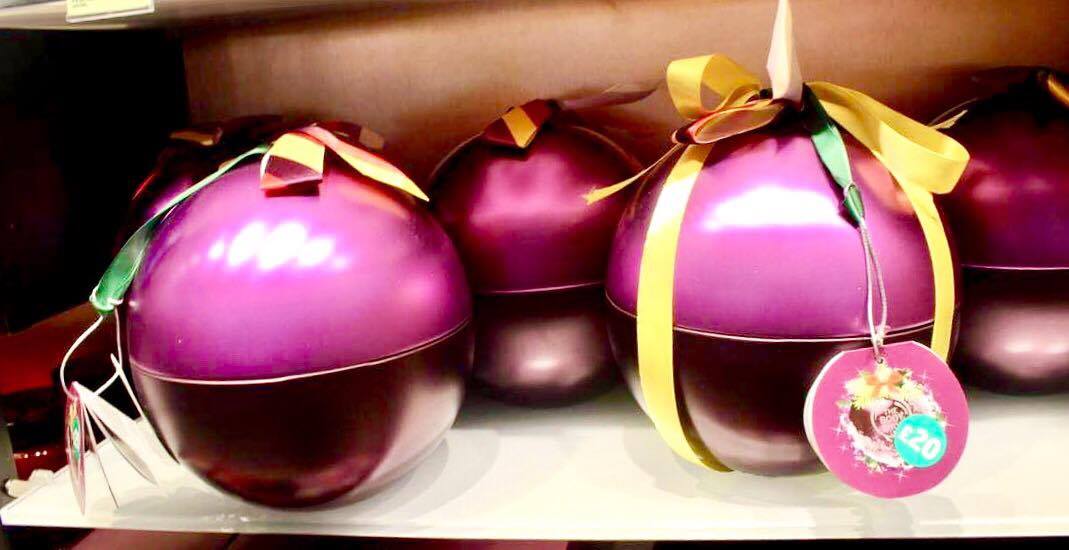Two large spherical metallic purple Plums with some bright green ribbon on top of them on a rectangular clear glass shelf, on a light background.