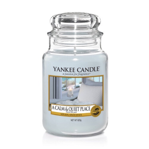 A tall glass jar full of some bright blue coloured wax with a label that has yankee candle written in black writing, a calm & quiet place written in black writing, and a picture of a kitchen with some white walls and blue countertops on it, on a bright background.