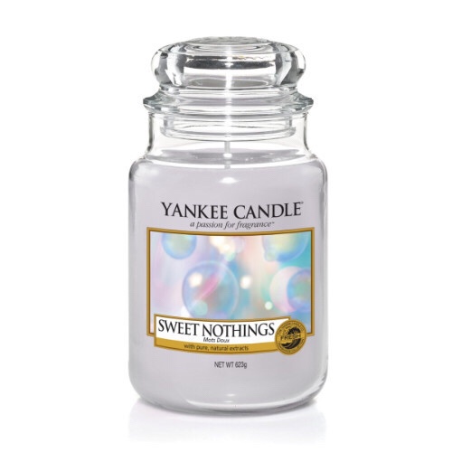 A tall glass jar full of some cream coloured wax with a label that has yankee candle written in black writing, sweet nothings written in black writing, and a picture of some white open flowers on a patch of green grass on it, on a bright background.