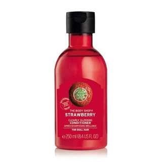 A tall cylindrical plastic bottle full of some bright cherry red liquid with a clear label that has a circular picture of a strawberry and The Body Shop Strawberry Clearly Glossing conditioner written in bold white writing on it, on a white background.
