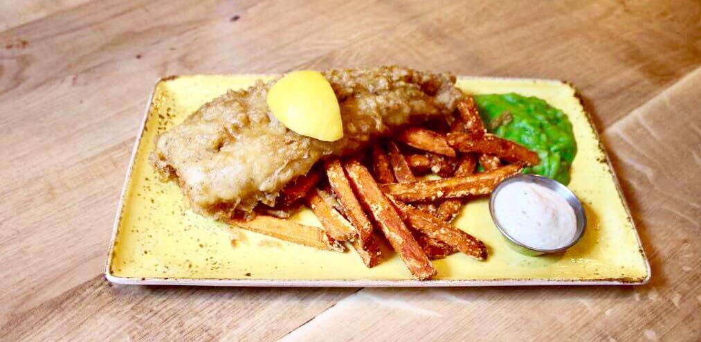 A long lightly battered cod fillet with a slice of Lemon on top of it next to some long rectangular sweet potato fries, a pile of light green mushy peas and a small white pot full of some creamy white tartar sauce on a white rectangular plate on a light wooden table, on a light background.