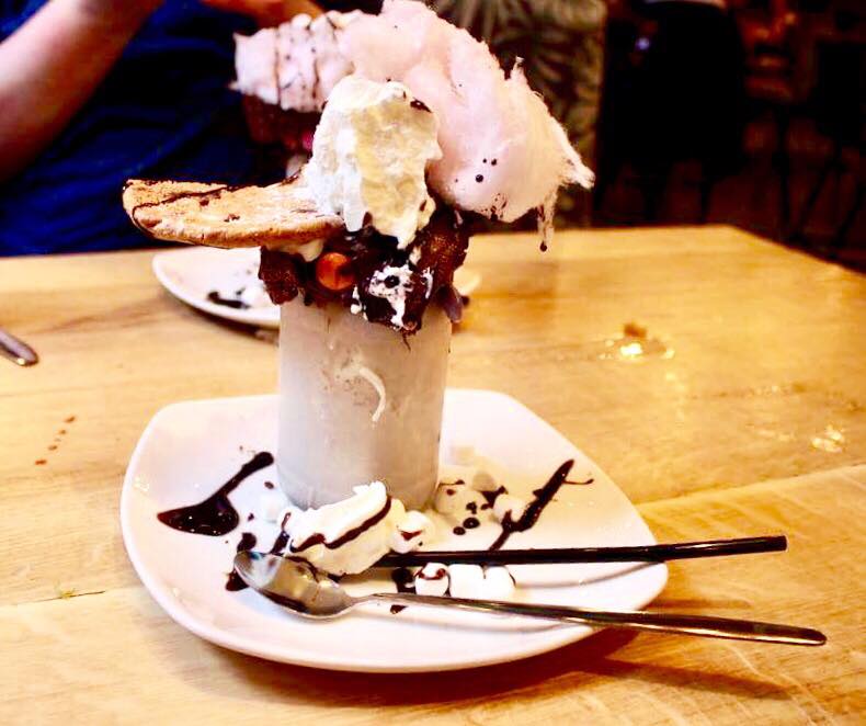 A tall milkshake glass full of some light brown liquid, some light brown chocolate fudge cake, a light brown chocolate chip cookie, some white squirty cream and some light pink candy floss on a white circular plate on a light wooden table, on a light background.