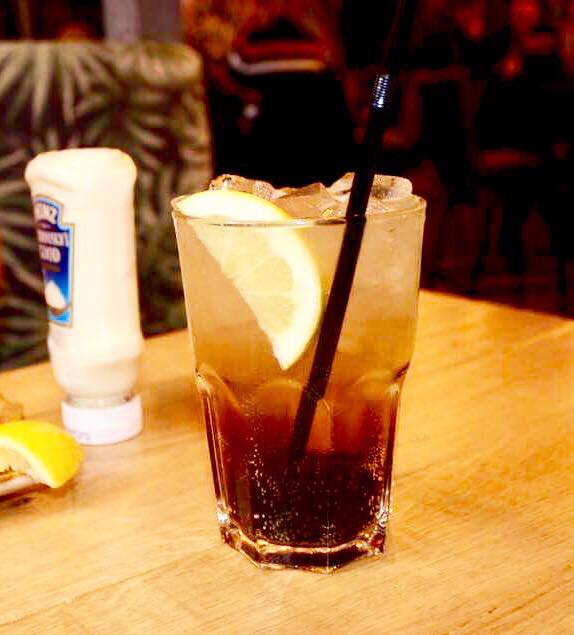 A tall high ball glass full of some dark brown liquid, some clear ice cubes, a slice of Lemon and a black straw on a light wooden table, on a light background.