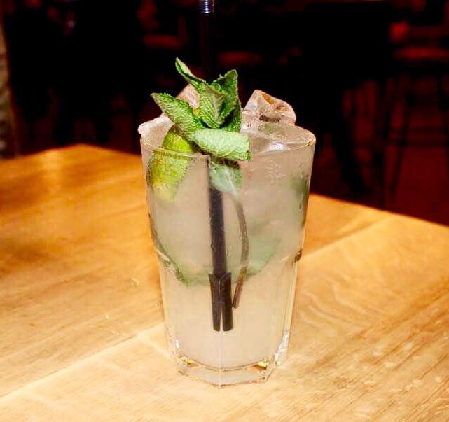 A tall high ball glass full of some clear liquid, some clear ice cubes, some mint leaves, some slices of Lime and a black straw on a light wooden table, on a light background.