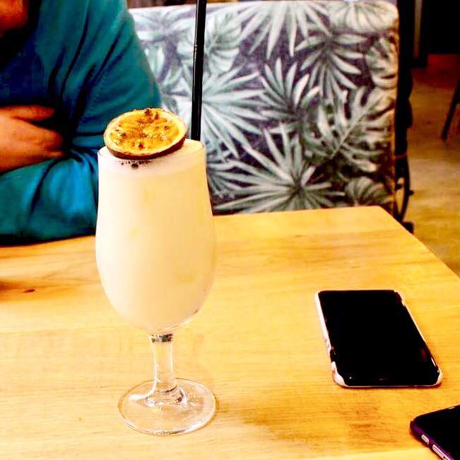 A tall high ball glass full of some white liquid, some clear ice cubes, half of a purple Passion Fruit and a black straw on a light wooden table, on a light floral background.