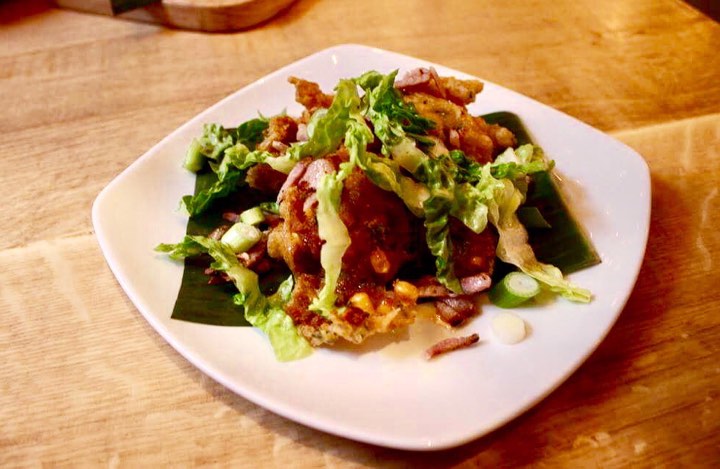Some golden brown balls with some yellow pieces of Sweetcorn, some dark brown pieces of Bacon, a light brown maple syrup glaze, some light green pieces of Spring Onion and some pieces of Red Chilli coming out of them on a rounded triangular plate on a light wooden table, on a light background.