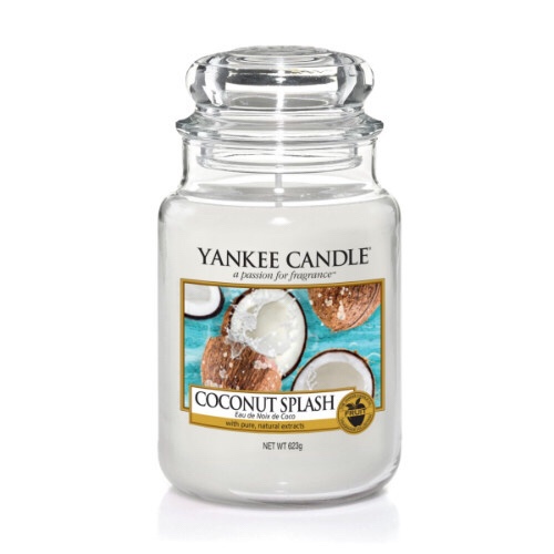 A tall glass jar full of some white coloured wax with a label that has yankee candle written in black writing, Coconut Splash written in black writing, and a picture of some grey coconut shells with creamy white insides next to some green spiky leaves on it, on a bright background.
