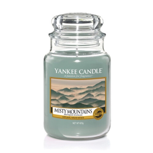 A tall glass jar full of some Ice Blue coloured wax with a label that has yankee candle written in black writing,Misty Mountains written in black writing, and a picture of a bright ice blue sky behind some ice white snow topped mountains on it, on a bright background.
