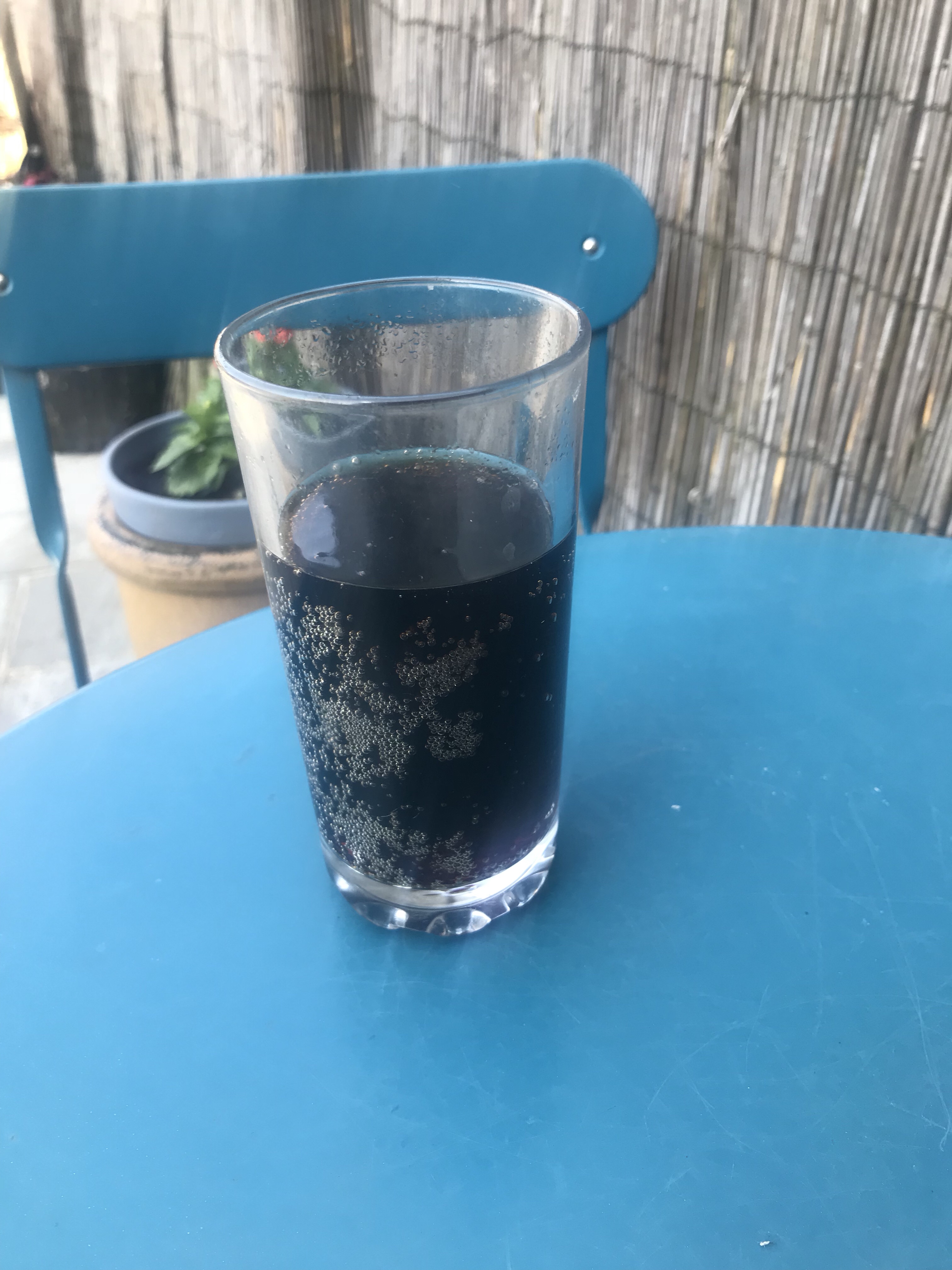 A clear glass full of some dark brown coloured liquid on top of a blue garden table, on a white background.