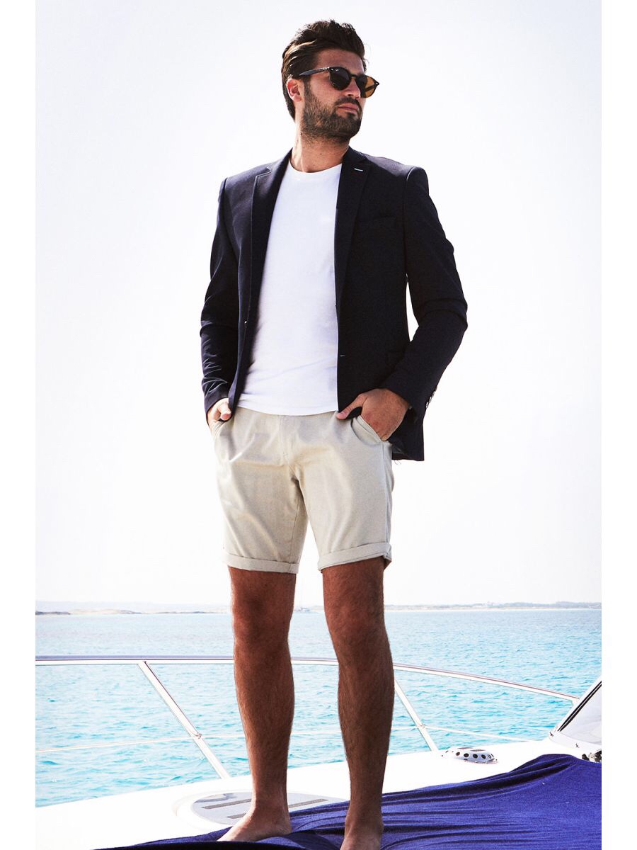 A man stood on a boat with the Sea in the distance wearing a pair of Beige Chino Shorts, a plain White T-Shirt, a navy blue blazer and a pair of dark Sunglasses, on a white background.
