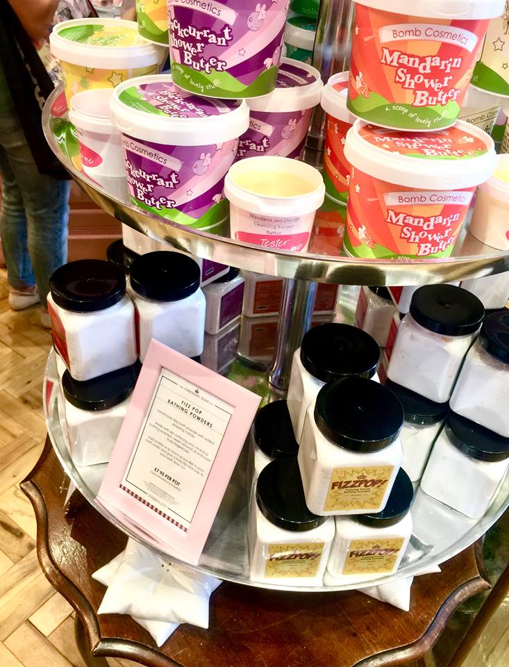 A tall circular clear glass two tiered table with some large cylindrical white pots on the top tier that have different coloured lids and labels that have Shower Butter written in large white writing on them, and some tall square plastic tubs on the bottom tier with black lids and labels that have Fizz Pop written in large red writing on them, on a bright background.
