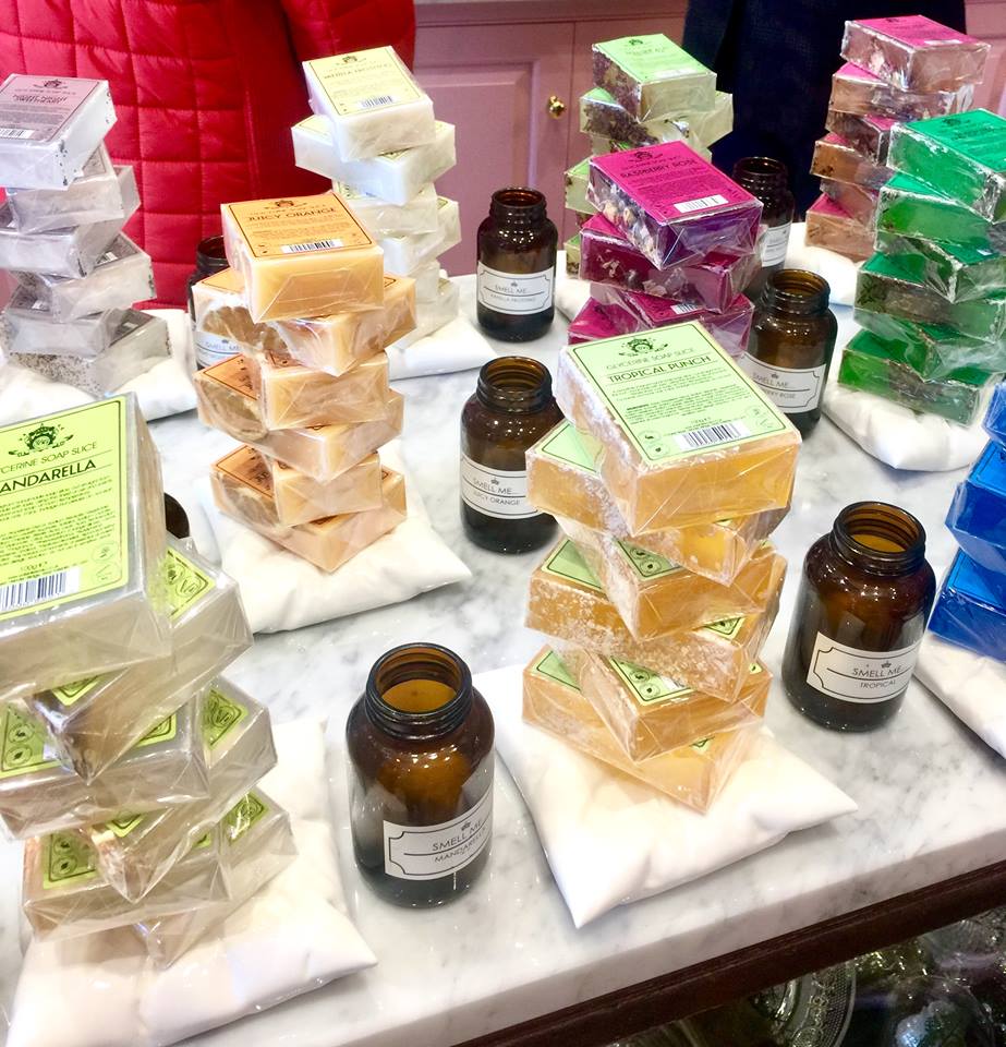 A long rectangular clear glass table that has some open black cylindrical pots and some square white marble slabs full of rectangular chunks of soap on it, on a bright background.