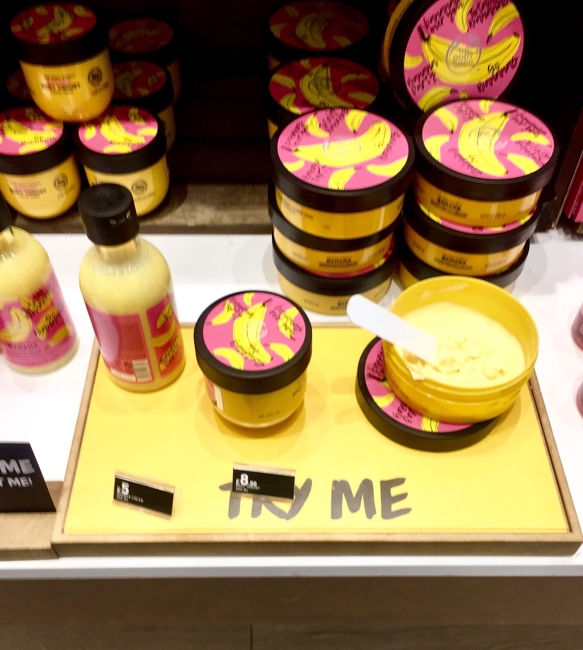 A tall cylindrical clear plastic bottle full of some yellow liquid with a label that has The Body Shop Banana Shower Cream written in bold black writing and some pictures of some Bananas on it, on a white background.