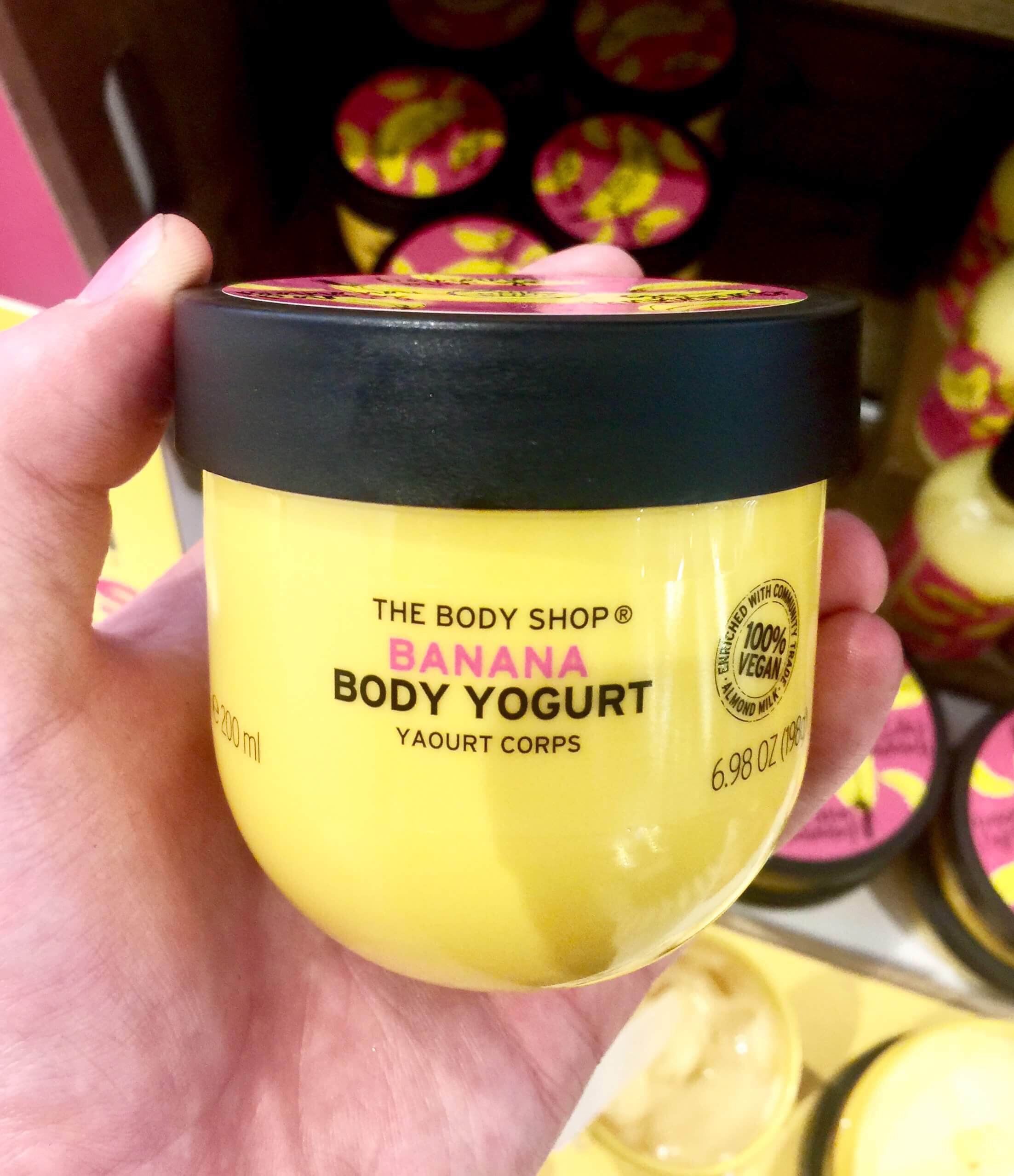 A short rounded square yellow plastic tub with a black lid that has The Body Shop Banana Body Yoghurt written in bold black writing and some pictures of some Bananas on it, on a white background.