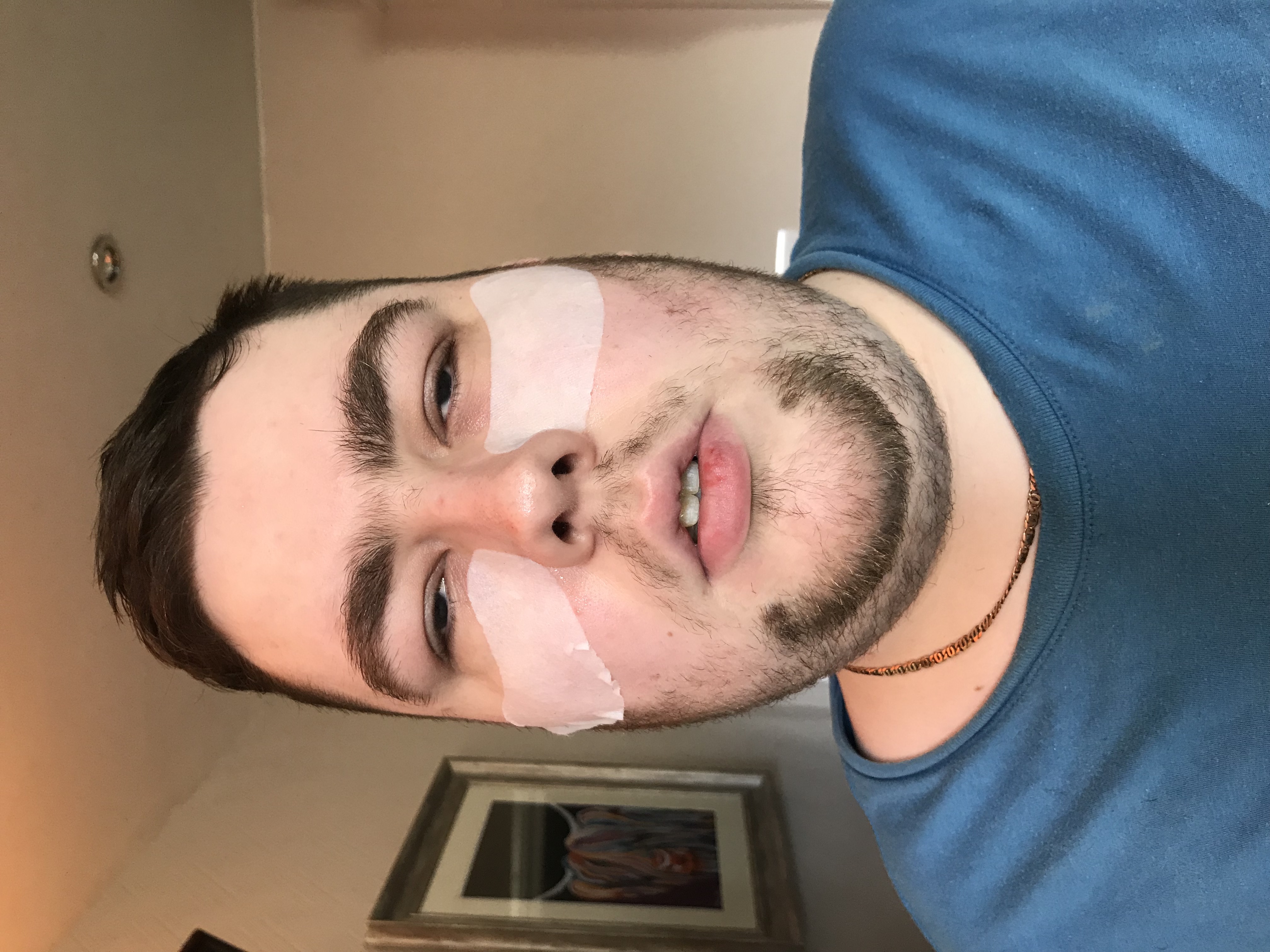 A bearded man wearing a green crew neck t-shirt and a piece of tissue underneath each eye, on a white background.
