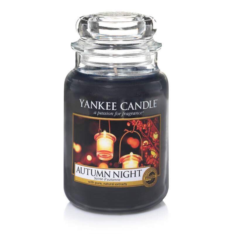 A tall glass Jar full of some dark Red coloured wax with a label that has yankee candle written in black writing, Autumn Night written in black writing, and a picture of some glass covered tea lights on top of some brown leaves on it, on a bright background.