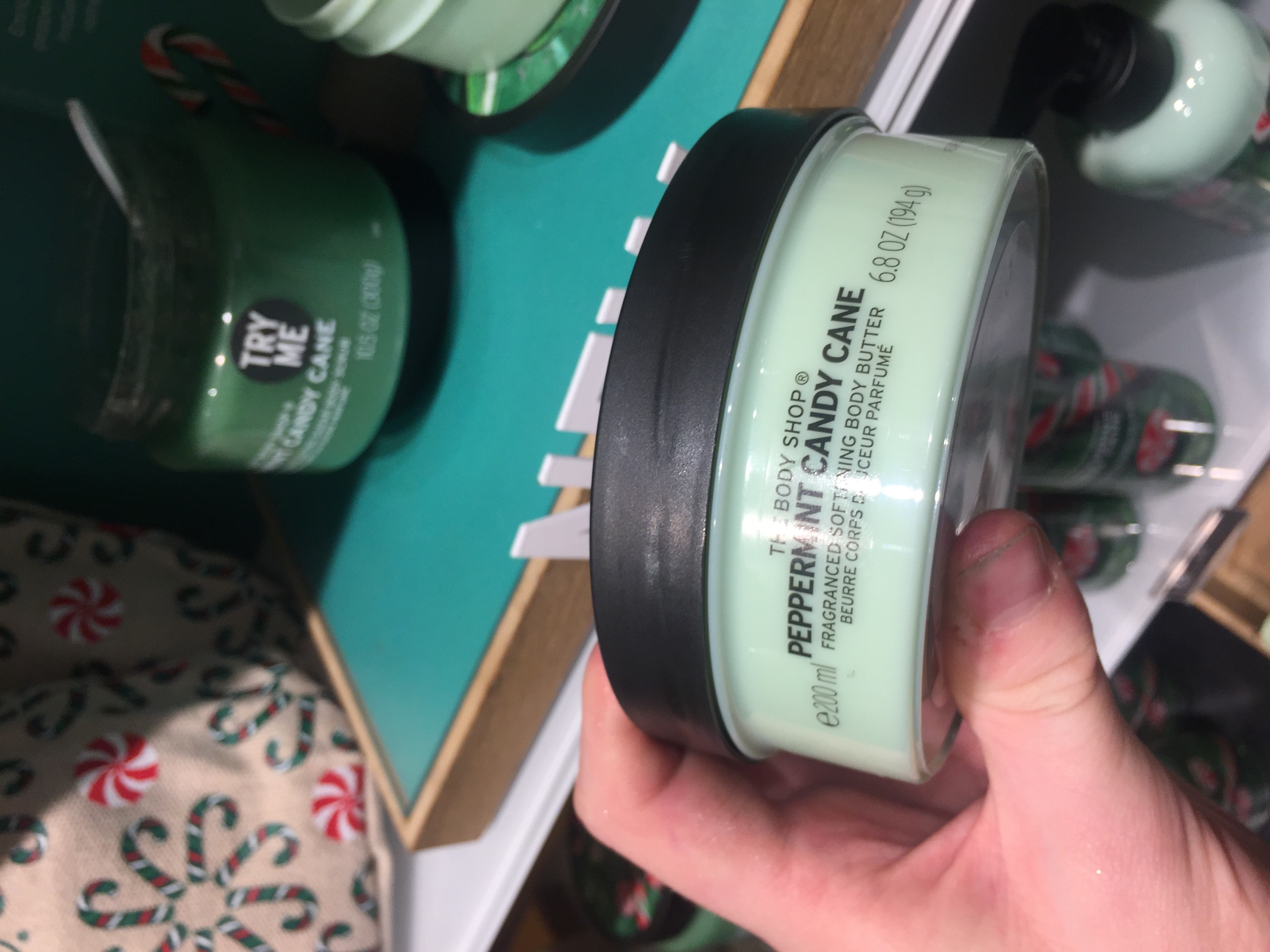 A short cylindrical white plastic tub with a green label that has The Body Shop Peppermint Candy Cane Body Butter written in bold black writing and some pictures of some red and white candy canes on it, on a white background.
