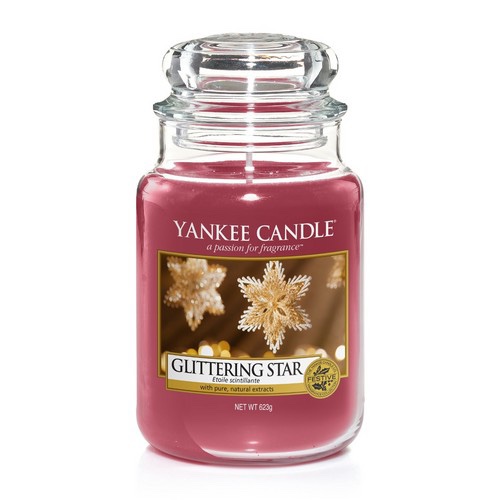 A tall cylindrical Glass Jar full of some dark Pink coloured Wax with a label that has Yankee Candle written in black writing, Glittering Star written in black writing, and a picture of some ornate Star shaped Baubles on it, on a white background.
