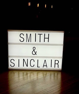 A large rectangular White Cardboard box that has Smith & Sinclair written in bold black writing on it on a large rectangular wooden table, on a dark background.