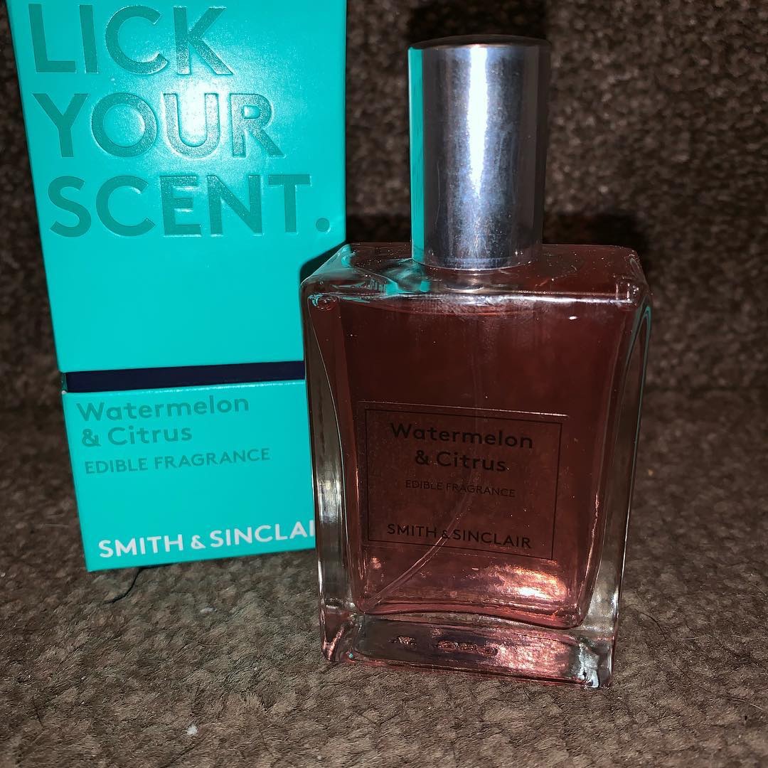 A tall rectangular clear glass bottle with a Silver metallic lid that has Smith & Sinclair Watermelon Citrus Edible Fragrance written in bold black writing on it next to a tall rectangular blue cardboard box that has Smith & Sinclair written in dark blue writing on it, on a white background.