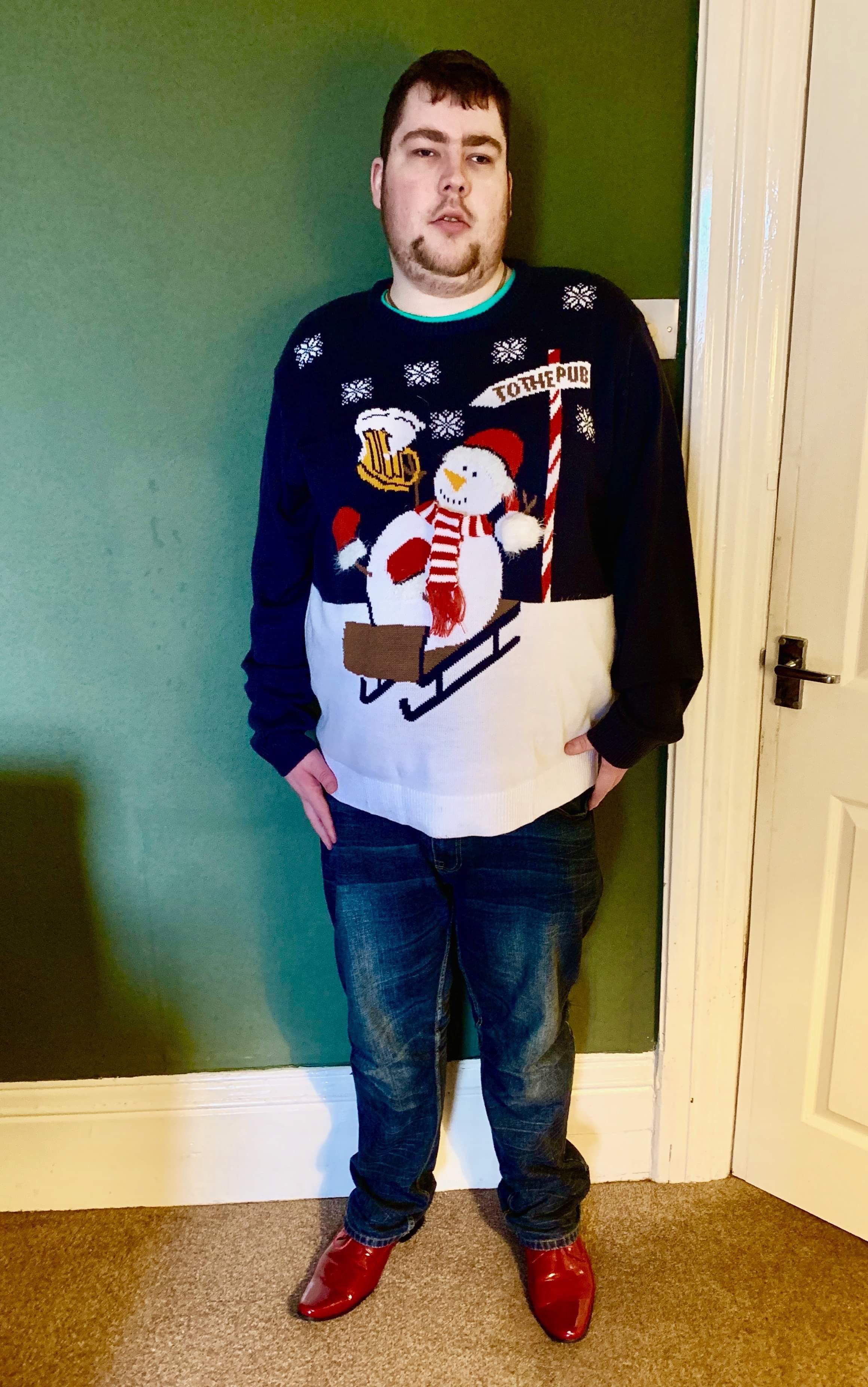 Me stood against a bright white wall wearing a navy jumper with a snowman design on it, a pair of navy jeans, and a pair of shiny red shoes, on a white background.