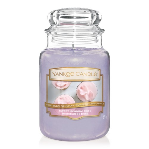 A tall Glass jar full of some bright pink wax with a label that has Yankee Candle written in gold writing, Sweet Morning Rose written in white writing, and a picture of three pink cupcakes on it, on a white background.