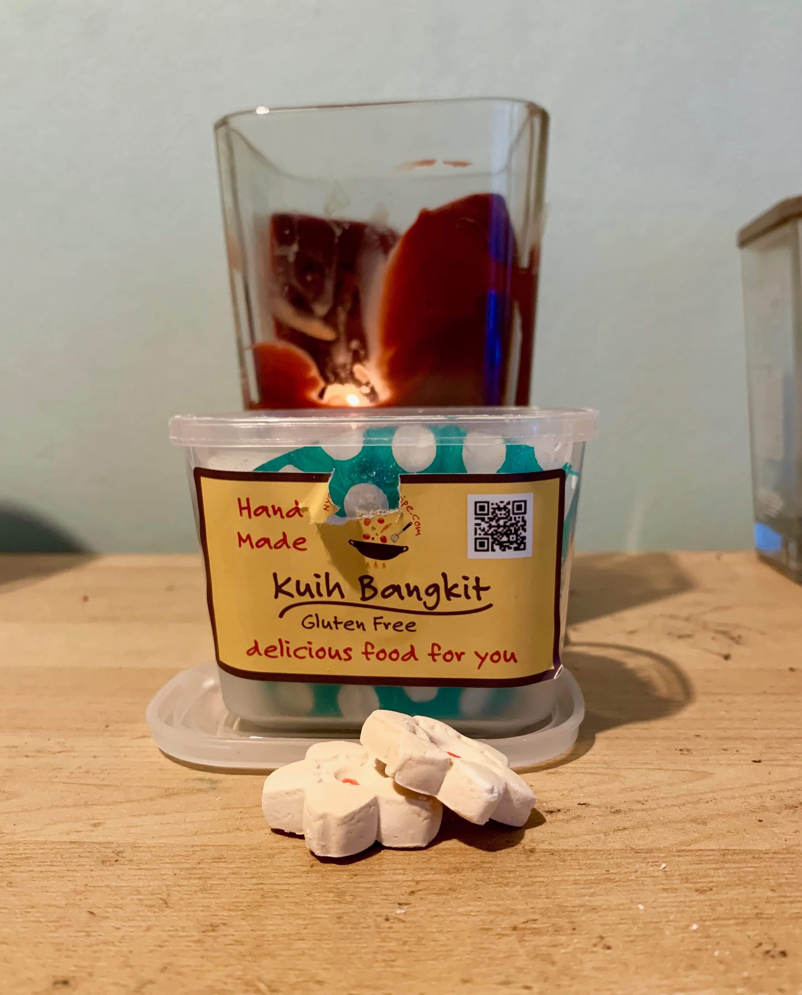 Some small star shaped light golden brown cookies in front of a short rectangular clear Plastic Tub with a yellow label that has nyonya recipe shop kuih bangkit cookies written in bold red writing on it on a light wooden table, on a white background.