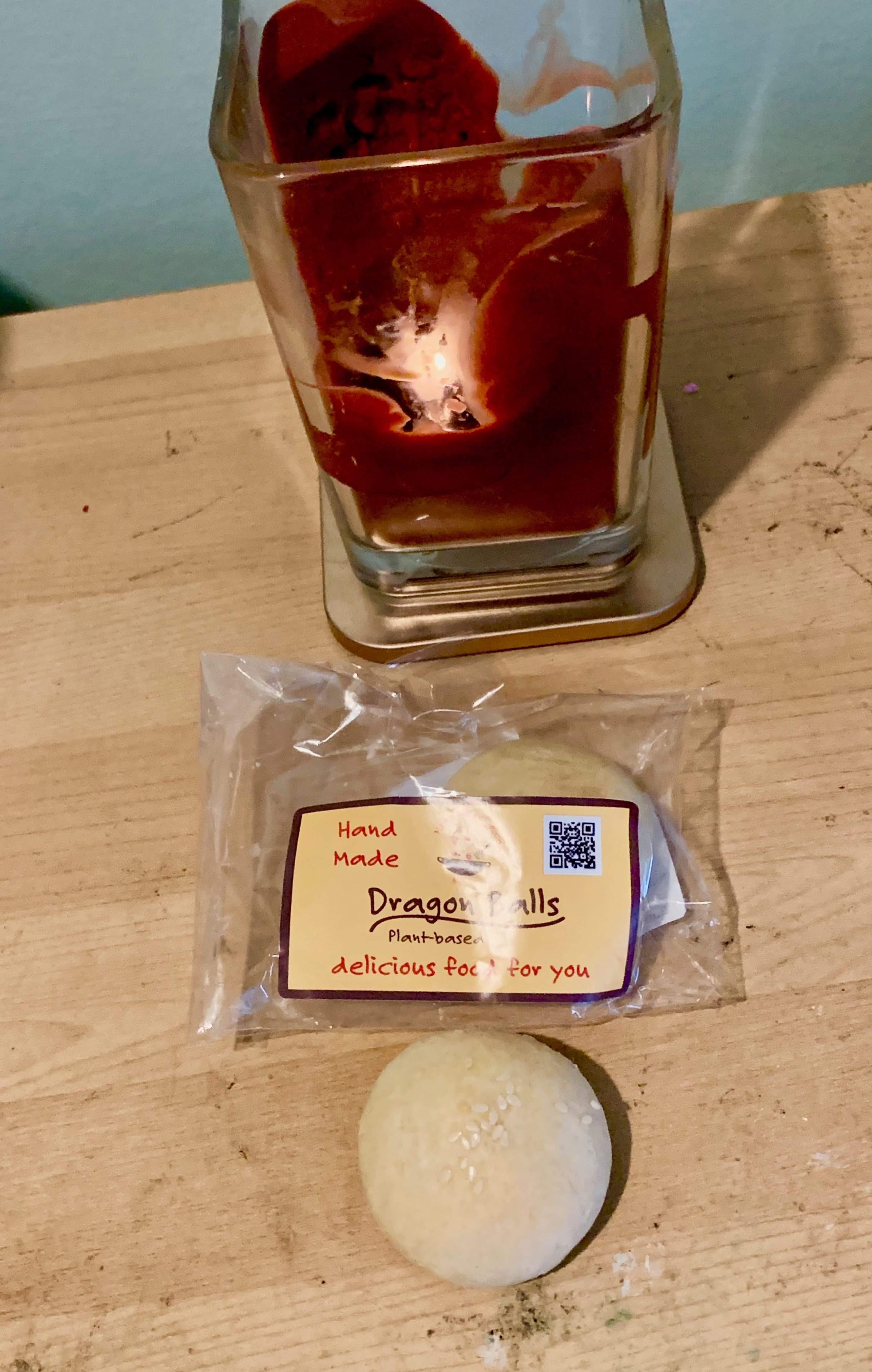A large circular dome of light golden brown pastry in front of a small rectangular clear Plastic Packet with a yellow label that has nyonya recipe shop dragon balls written in bold red writing on it on a light wooden table, on a white background.