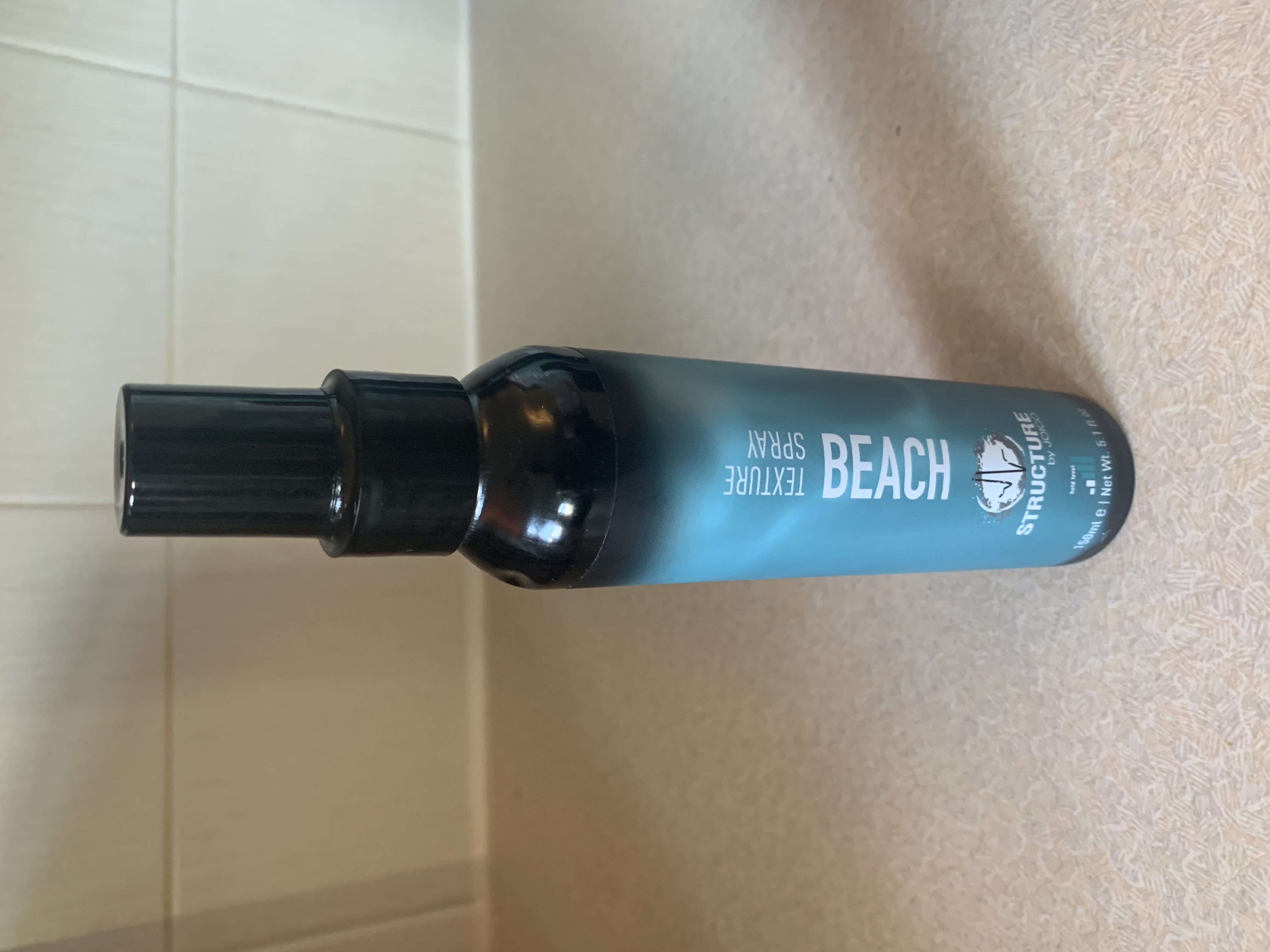 a tall cylindrical Black Plastic spray bottle with a white label that has Structure by Joico Beach written in dark blue writing on it, on a white background.